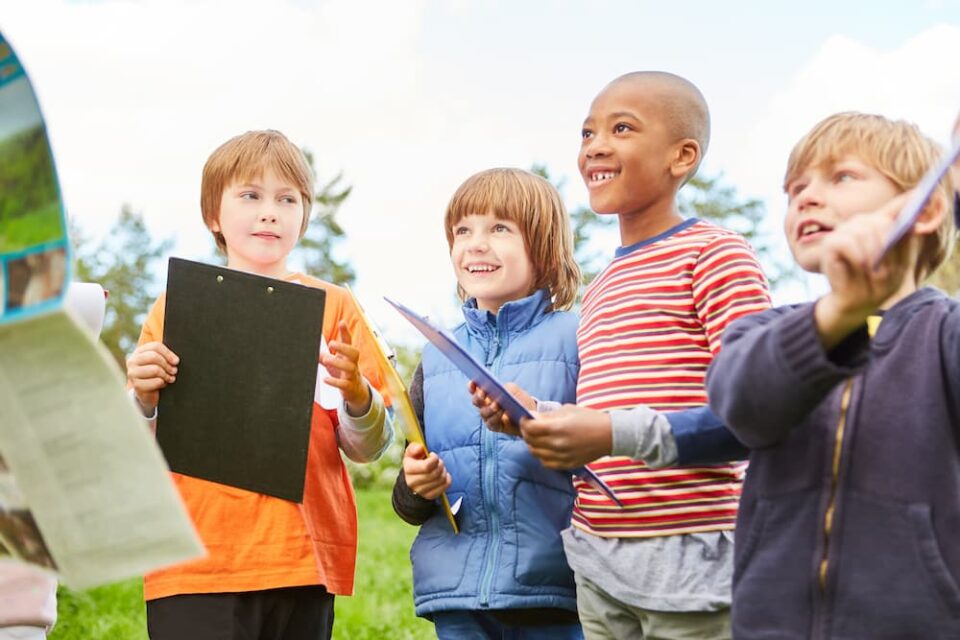 Four kids outside in a backyard holding clipboards, receiving instructions for a scavenger hunt