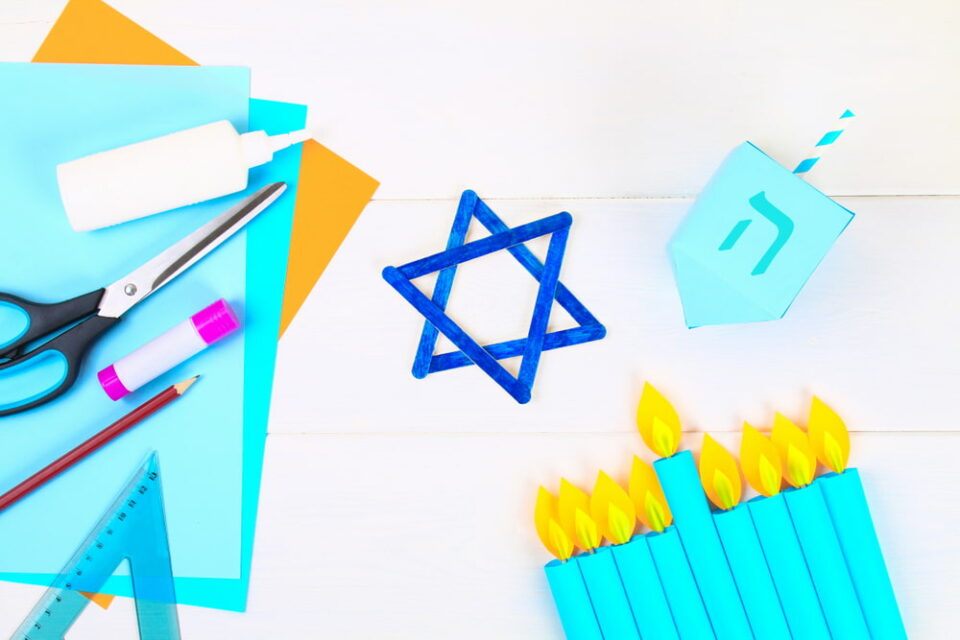 A popsicle stick Star of David, a paper dreidel, and a paper menorah on a white table.
