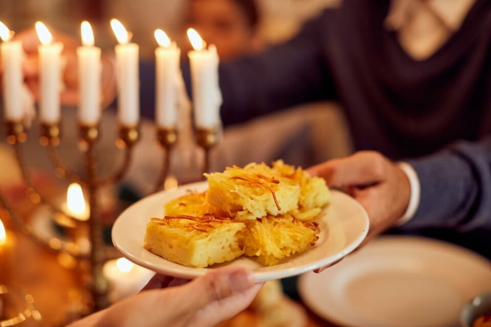 Hands passing a plate of kugel across a table with a menorah in the background
