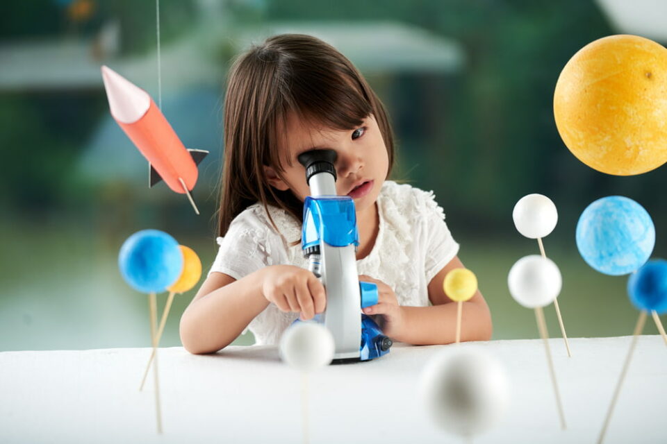 A curious girl surrounded by model planets looking through a toy microscope.