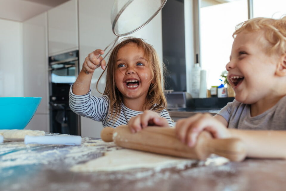 A smiling little girl holding a strainer with a boy making dough using a rolling pin on the kitchen table