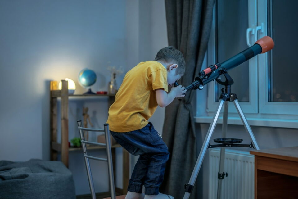 A-young-boy-is-looking-through-a-telescope-in-his-bedroom-at-the-starry-night-sky