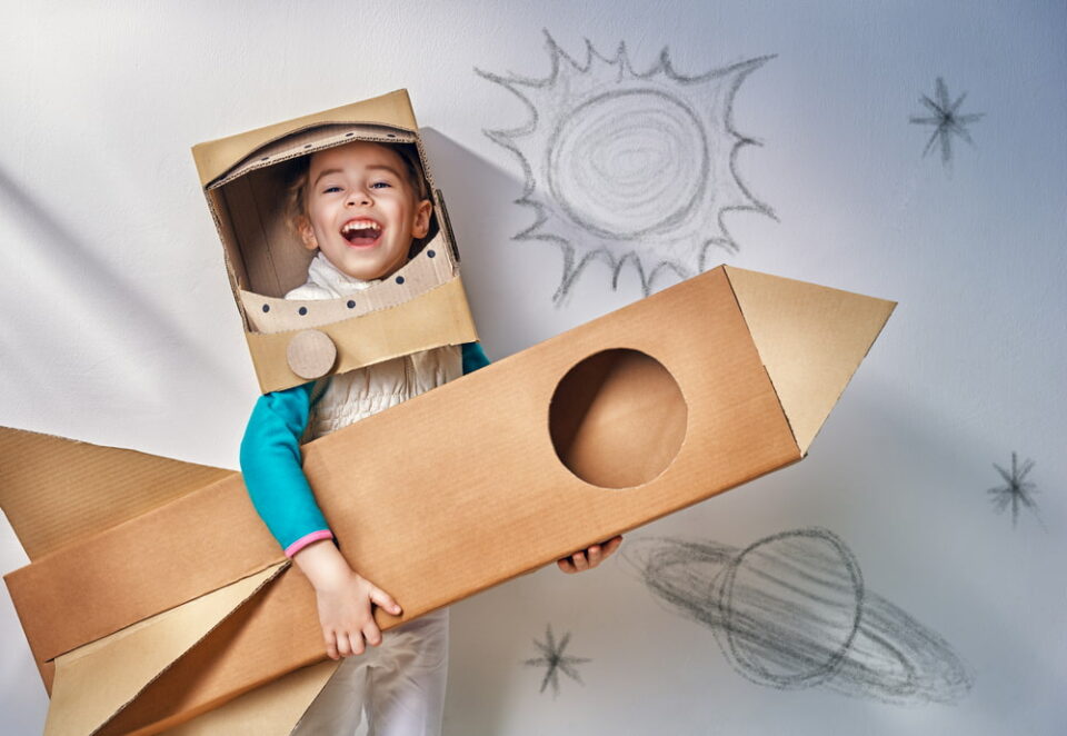 A-young-boy-laughing-and-holding-a-large-cardboard-rocket-and-wearing-a-cardboard-helmet