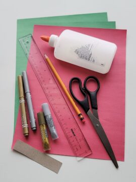 Glue, scissors, pencil, markers, cardboard, glitter glue, and ruler on top of construction paper