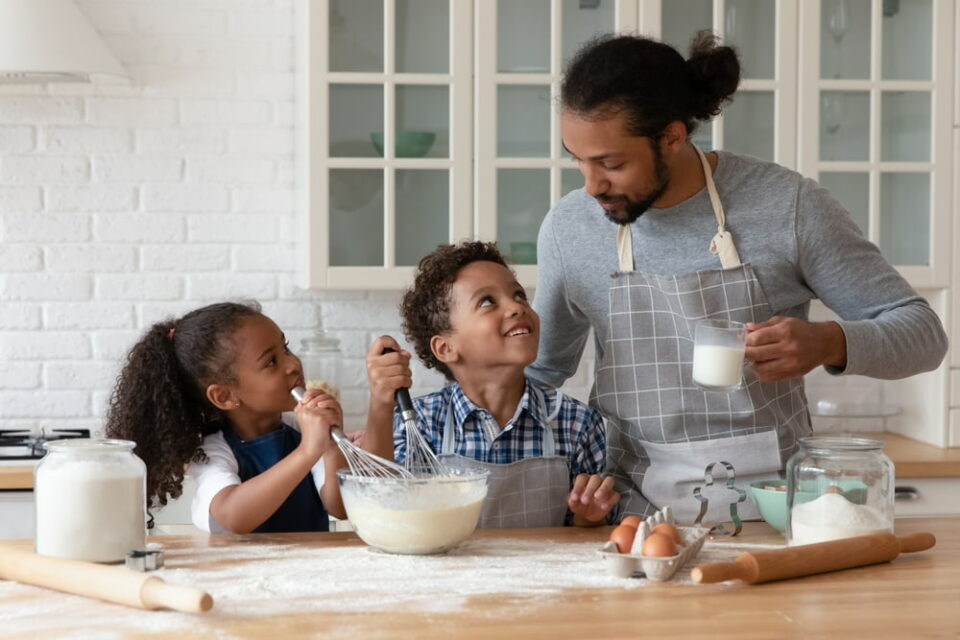 Kids and a dad wearing aprons in a kitchen mixing eggs, milk, and flour to make cookie batter