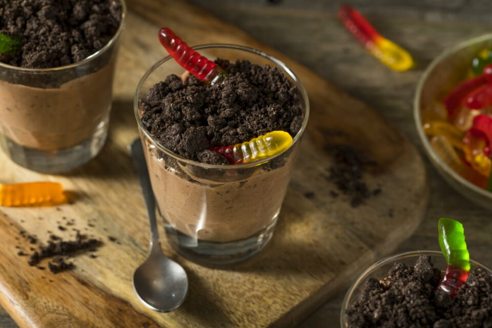 Dirt and worm pudding cups arranged on a wooden board. The cups are garnished with colorful gummy worms