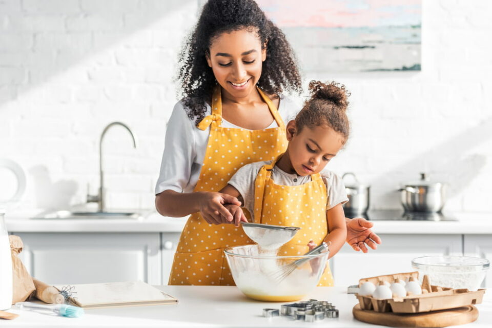 A young girl with her mother sieving flour into a batter in the kitchen.