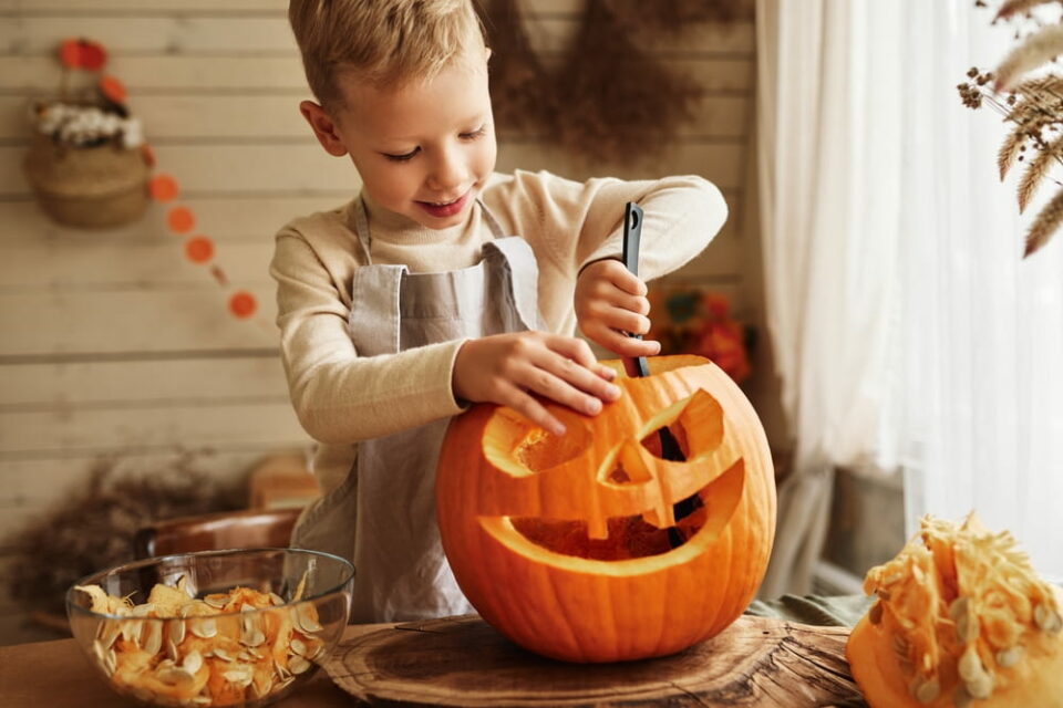 A young boy in an apron scooping out the insides of a large, carved jack-o’-lantern pumpkin