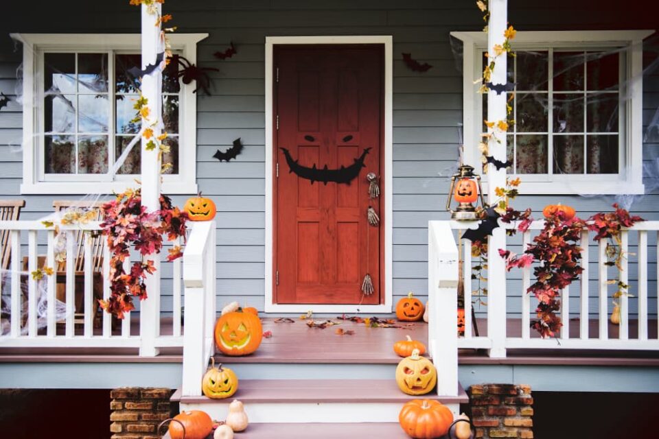 A front porch is decorated for Halloween with pumpkins, leaves, bats, and a spooky face on the front door