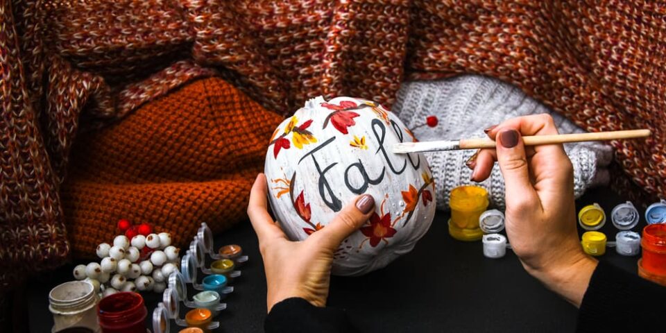 A woman holding a brush to a painted pumpkin that reads “Fall.”