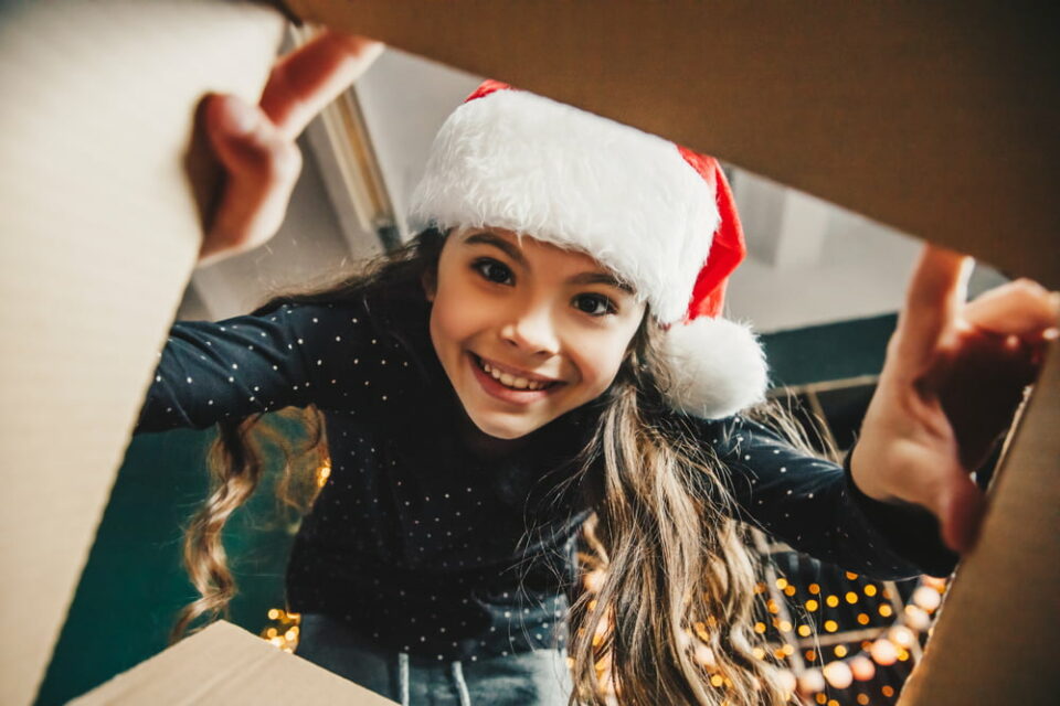 A girl wearing a Santa hat, smiling and looking inside a box
