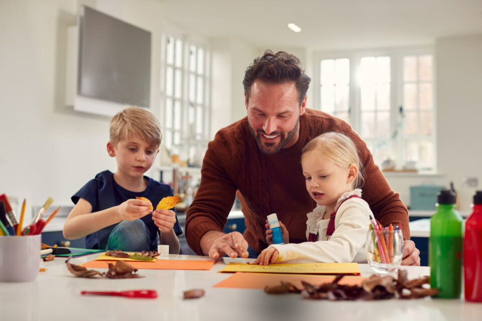 A father and two young children sit at the kitchen counter creating fall crafts using leaves.