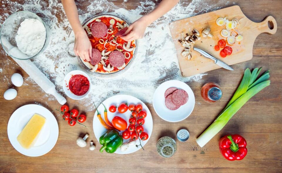 A table covered in flour and ingredients with hands adding pepperoni to a pizza.