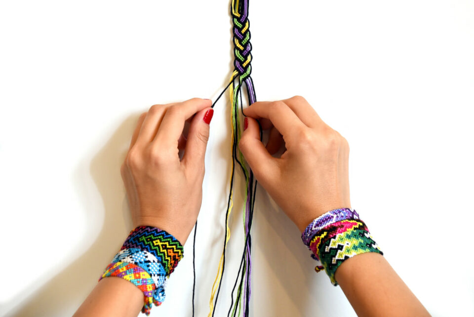 Two hands, wrists adorned with friendship bracelets, tie a bracelet using colorful threads.
