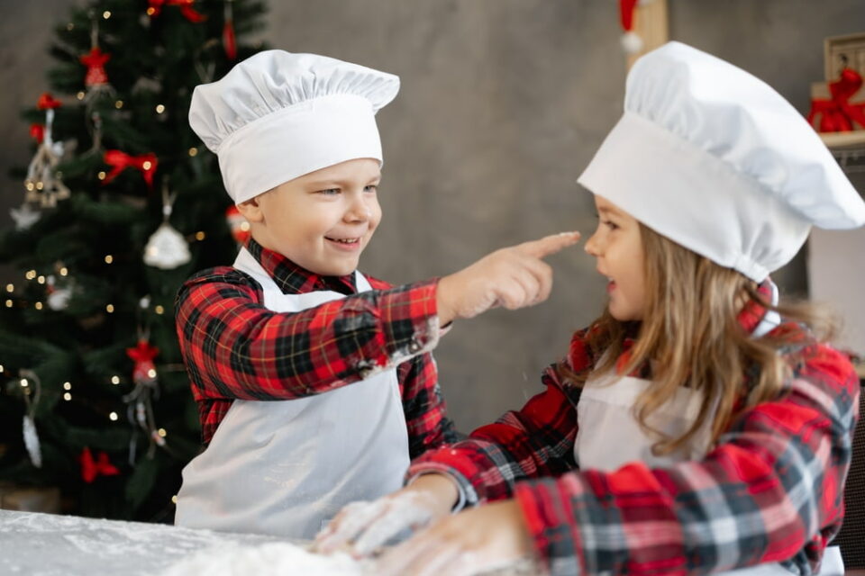 Two young children bake together with a Christmas tree in the background