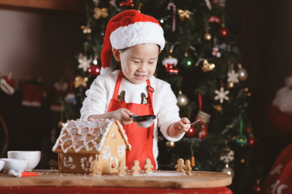 A young boy wearing a Santa hat decorates a gingerbread house with icing.