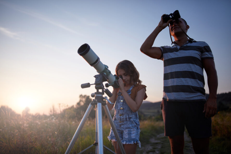A father and daughter outside looking through a telescope at sunset