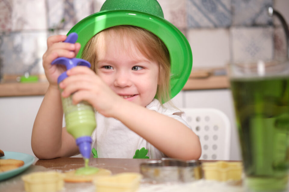 A child in a green hat, icing a cookie with green frosting.