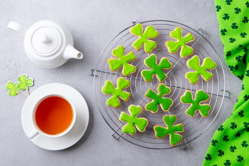 Shamrock-shaped cookies with green frosting on a cooling rack next to a pot of tea.