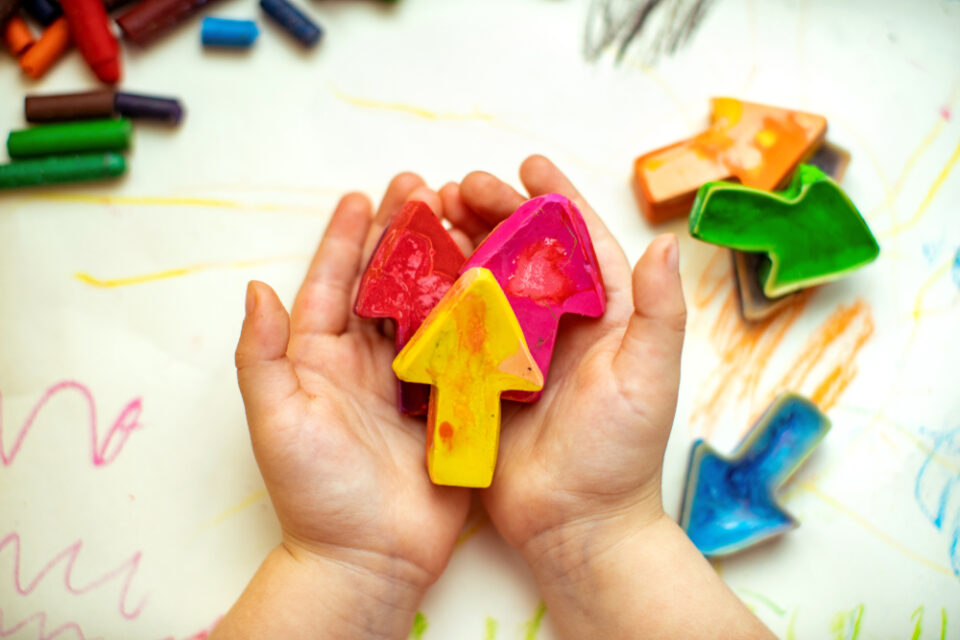 A child’s hands holding yellow, red, and pink crayon arrows, with other assorted crayons on the table below.