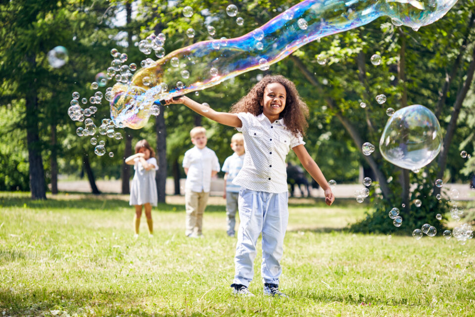 A girl making a giant bubble outside with three other kids standing behind her.