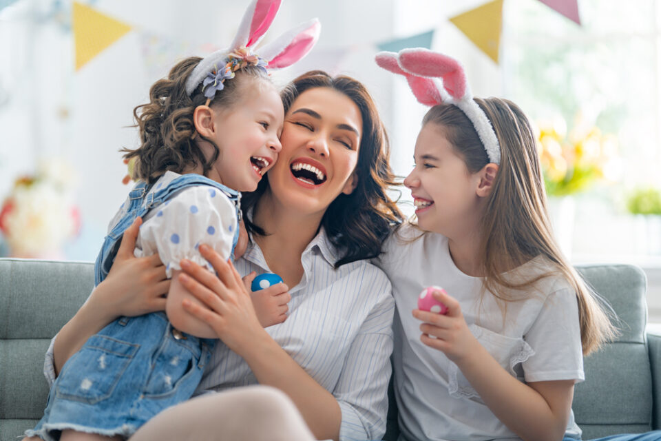 A mom and two girls wearing bunny ears, laughing together and holding Easter eggs.