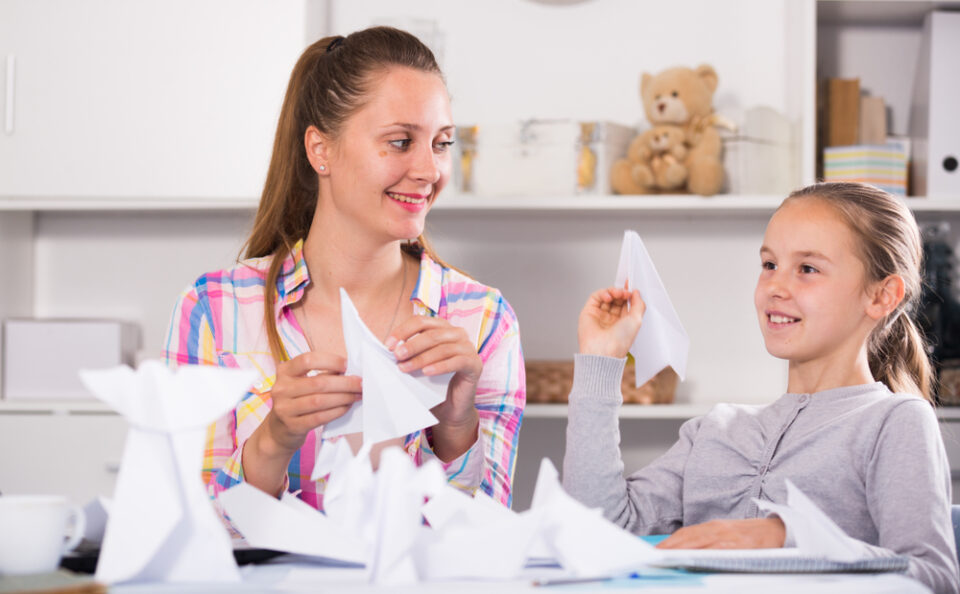 A mother and daughter folding paper airplanes at a table.