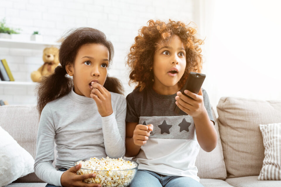 Two kids watching TV at home and eating popcorn.