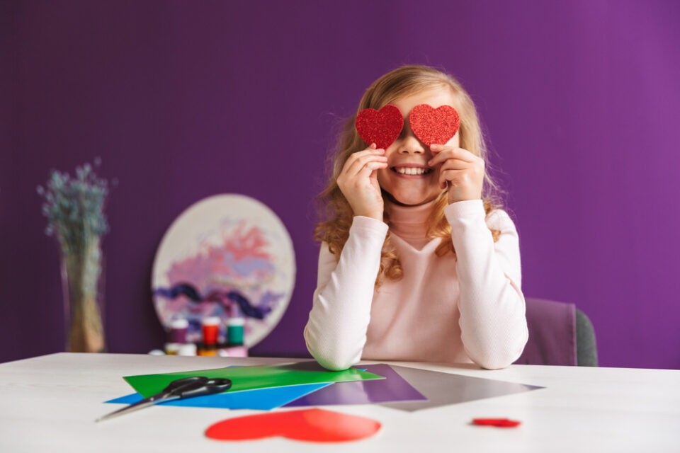 A girl sitting at a table with craft paper holding two red sparkly hearts over her eyes.