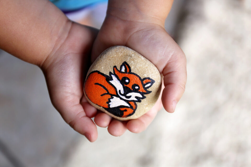 A child’s hands holding a rock painted to look like a rainbow.