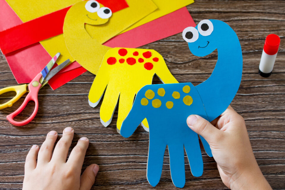 A child’s hands holding a blue handprint dinosaur. A yellow handprint dinosaur sits on the table behind it