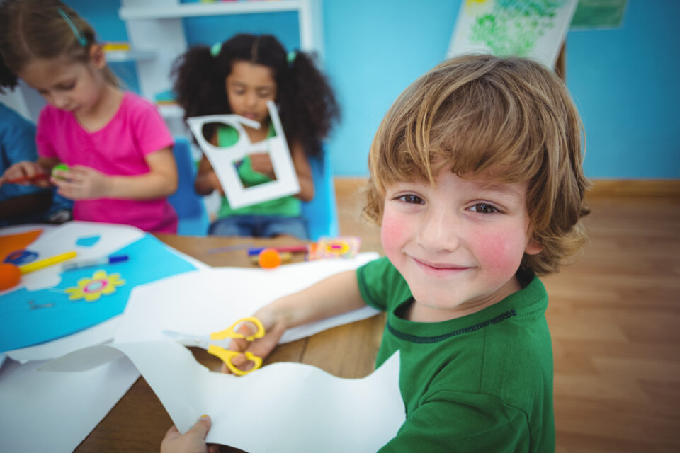 A little boy smiling as he cuts a piece of white paper for a craft