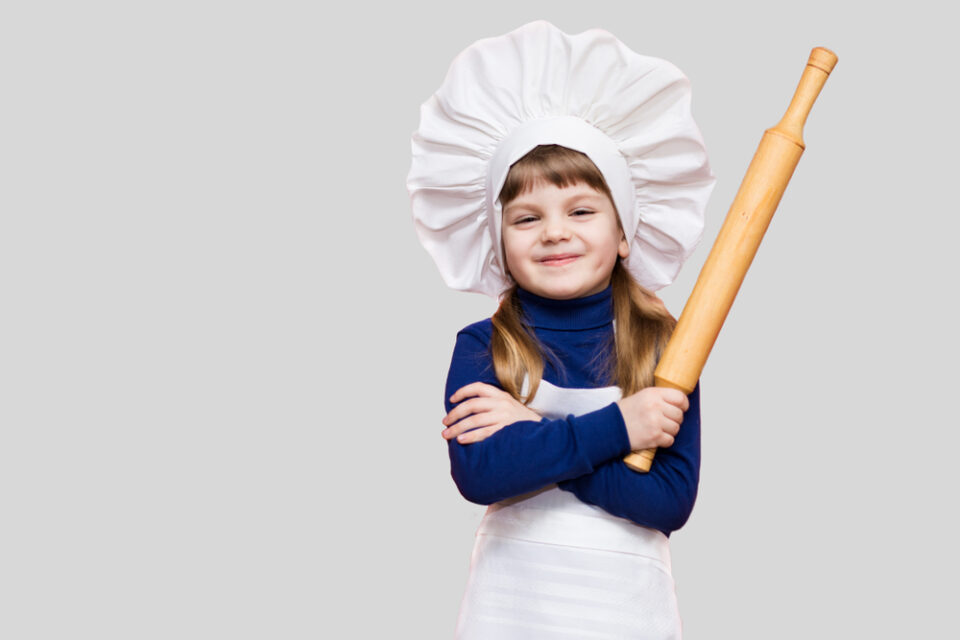 A smiling girl holding a rolling pin and wearing a chef’s hat and apron.