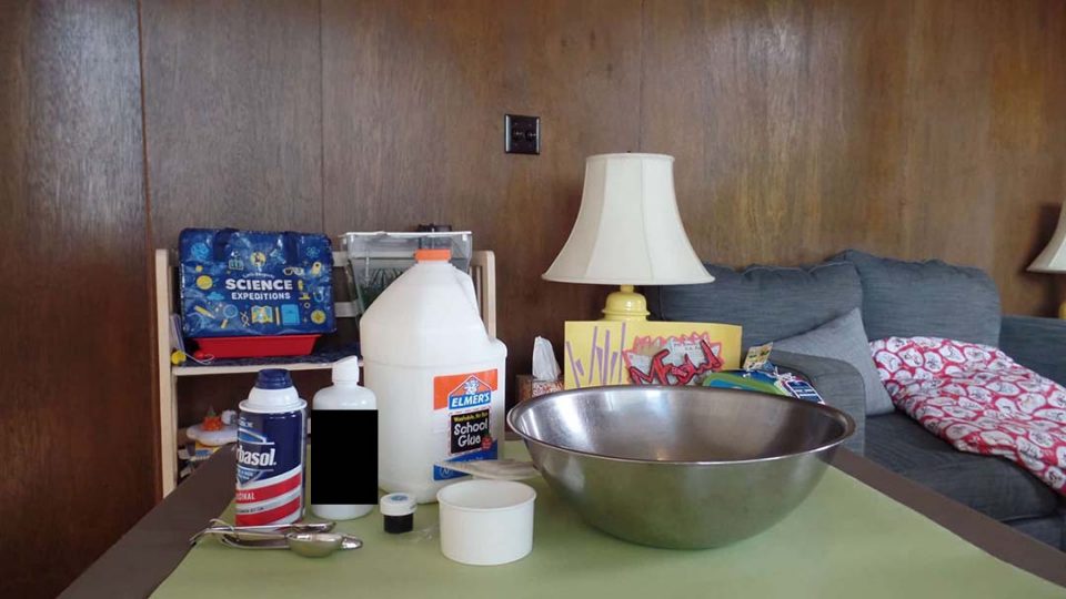 Fluffy slime ingredients: glue, shaving cream, a bowl, and food coloring on a table